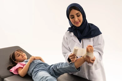 ايماءات السعادة و الارتياح بعد تلقي العلاج المناسب , طفلة خليجية عربية سعودية ترتدي الزي الكاجوال نائمة على سرير الكشف تشعر بالراحة عند وضع الضمادة الطبية , طبيبة عربية خليجية سعودية محجبة ترتدي المعطف الطبي الابيض تقدم المساعدة اللازمة للفتاة المصابة , بورتريه مقرب , خلفية بيضاء