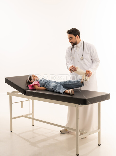 A nice friendly conversation between the doctor and the girl while receiving appropriate treatment, an Arab Gulf Saudi doctor wearing a white medical coat is putting a bandage on the little girl, an Arab Gulf Saudi girl wearing casual clothes is lying on the examination bed while her foot is being examined, portrait with a white background, full body.