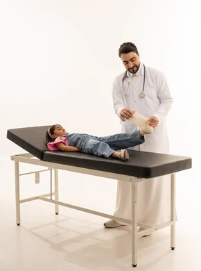 Providing appropriate assistance for each medical case with high efficiency, an Arab Gulf Saudi doctor wearing a white medical coat is applying a medical bandage to a young girl, an Arab Gulf Saudi girl dressed in casual attire raises her foot to receive the necessary treatment, portrait with a white background, full body.