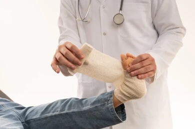 بورتريه مقرب لطبيب هيئة الهلال الاحمر السعودي يمارس العمل باحترافية تامة , طفلة عربية خليجية سعودية ترتدي الزي الكاجوال نائمة على سرير الكشف و ترفع القدم المتألمة , طبيب عربي خليجي سعودي يرتدي المعطف الطبي و يضع الضمادة الطبية للفتاة الصغيرة , خلفية بيضاء