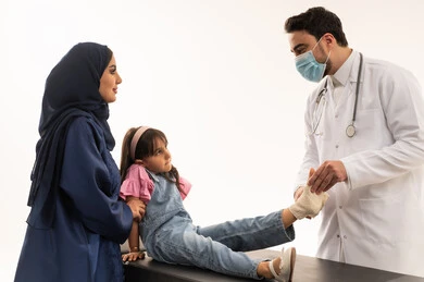 طبيبة عربية سعودية خليجية محجبة ترتدي العباءة الزرقاء تساعد البنت الصغيرة اثناء وضع الضمادة الطبية , طبيب عربي خليجي سعودي يرتدي الكمامة للحدمن انتشار العدوى , طفلة عربية خليجية سعودية ترتدي الزي الكاجوال تجلس على سرير الكشف , بورتريه مقرب , خلفية بيضاء