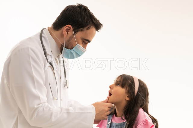استخدام الكمامة يساعد على تقليل انتشار الامراض المعدية , طبيب عربي خليجى سعودي يرتدي الكمامة و المعطف الطبي الابيض يفحص اسنان الفتاة الصغيرة للكشف عن التسوس , طفلة عربية خليجية سعودية ترتدي الزي الكاجوال تجلسعلى سرير الكشف الاسفنجي , بورتريه مقرب , خلفية بيضاء