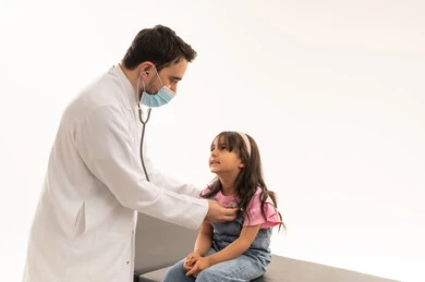 حماية النفس و الغير من الامراض مع اتمام الفحص المطلوب  , طبيب عربي خليجي سعودي يرتدي الكمامة و معطف طبي ابيض يستمع الي قلب الصغيرة , فتاة عربية خليجية سعودية ترتدي الزي الكاجوال جالسة على سرير الكشف , بورتريه مقرب , خلفية بيضاء