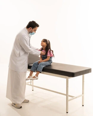 نظرة للطبيب مليئة بالامل في الشفاء , طفلة عربية خليجية سعودية ترتدي الزي الكاجوال تجلس على سرير الكشف , طبيب عربي خليجي سعودي يرتدي الكمامة و المعطف الطبي الابيض يستمع لنبضات قلب الصغيرة , بورتريه خلفية بيضاء , الجسم بالطول الكامل