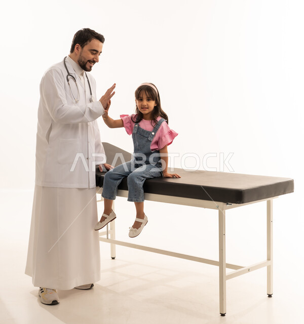 النظر للكاميرا مع ايماءات السعادة للانتصار على الامراض , بنت صغيرة عربية خليجية سعودية تجلس على سرير الكشف ترتدي الزي الكاجوال , طبيب عربي خليجي سعودي يرتدي المعطف الطبي الابيض يصافح بالكفين عالياً الطفلة بعد انتهاء الفحص الطبي , بورتريه خلفية بيضاء ,الجسم بالطول الكامل