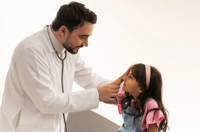 العناية بالعيون في هيئة الهلال الاحمر السعودي , طبيب عربي خليجي سعودي يرتدي معطف طبي ابيض يقوم بفحص عين الطفلة , طفلة عربية خليجية سعودية ترتدي الزي الكاجوال تجلس على سرير الكشف لاتمام فحص العيون , بورتريه مقرب , خلفية بيضاء