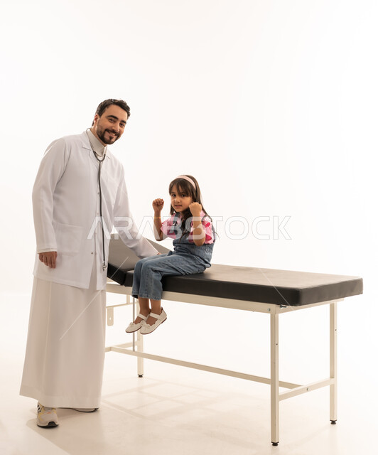 بورتريه خلفية بيضاء ,الجسم بالطول الكامل ,طفلة خليجية عربية سعودية تجلس على سرير الكشف في العيادة وترفع اليدين علامة على الفرحة بانتهاء الرحلة العلاجية ,طبيب عربي خليجي سعودي يرتدي معطف طبي ابيض يقف بجانب الطفلة ,ايماءات السعادة و السرور ,النظر للكاميرا