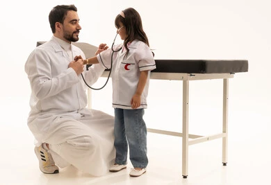 Moments of affection and happiness between the doctor and the girl, a Saudi Gulf Arab girl wearing a doctor's costume and examining the doctor while listening to his heartbeat. An Arab Gulf Saudi doctor wearing a white medical coat squats in front of the girl and pretends to be the patient, with gestures of smiles and joy, portrait with a white background, full-body.