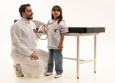 طفلة عربية خليجية سعودية صغيرة تتقمص دور الطبيب ترتدي زي تنكري تنظر للكاميرا و تلعب مع طبيبها بمرح , طبيب عربي خليجي سعودي يرتدي معطف طبي ابيض يجلس القرفصاء امام الطفلة و ينظر لها في حنان ,بورتريه خلفية بيضاء,الجسم بالطول الكامل ,