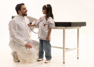 طبيب عربي سعودي خليجي يرتدي المعطف الطبي الابيض ينظر الى الفتاة الصغيرة وهو يتقمص دور المريض ,طفلة عربية خليجية سعودية ترتدي زي طبيب تنكري و تحمل سماعة الطبيب و تسمع نبضات القلب ,ايماءات بهجة وسرور, بورتريه خلفية بيضاء ,الجسم بالطول الكامل