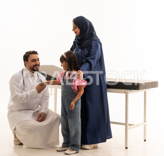 الابقاء على العلاقة الودودة بين المرضى و الاطباء مع الحفاظ على الجدية في أداء المهنة ,طبيب عربي خليجي سعودي يرتدي المعطف الطبي الابيض يجلس القرفصاء امام الطفلة الصغيرة ,طبيبة عربية خليجية سعودية محجبة ترتدي عباءة زرقاء تمسك بالطفلة الصغيرة بعد الانتهاء من الفحص , بورتريه خلفية بيضاء , الجسم بالطول الكامل