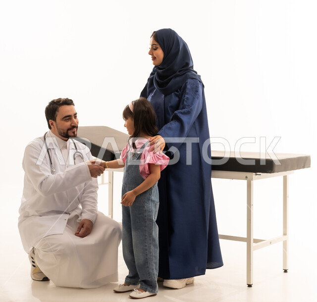 طبيبة عربية خليجية سعودية محجبة ترتدي عباءة زرقاء تقف بجوار الطفلة لمساعدتها ,طبيب عربي خليجي سعودي يرتدي المعطف الطبي الابيض يجلس القرفصاء امام طفلة صغيرة بابتسامة بعد استقرار الحالة و الاطمئنان على الصحة , بورتريه خلفية بيضاء , الجسم بالطول الكامل