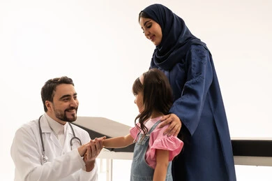 طبيب عربي خليجي سعودي يرتدي المعطف الطبي الابيض يجلس القرفصاء امام الطفلة بعد الانتهاء من الفحص , طبيبة عربية خليجية سعودية محجبة ترتدي عباءة زرقاء تقف بجانب الطفلة بعد الاطمئنان من الفحص , بورتريه مقرب ,خلفية بيضاء