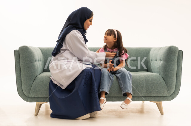 مزاولة مهنة الطب بجدية , طفلة عربية خليجية سعودية ترتدي الزي الكاجوال تجلس بجانب الطبب لاستكمال الفحوصات الضرورية , طبيبة عربية خليجية سعودية محجبة ترتدي المعطف الطبي الابيض تستمع بانصات لنبضات قلب الصغيرة للتأكد من سلامتها , بورتريه الجسم بالطول الكامل , خلفية بيضاء