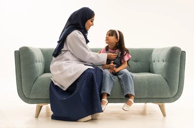 مزاولة مهنة الطب بجدية , طفلة عربية خليجية سعودية ترتدي الزي الكاجوال تجلس بجانب الطبب لاستكمال الفحوصات الضرورية , طبيبة عربية خليجية سعودية محجبة ترتدي المعطف الطبي الابيض تستمع بانصات لنبضات قلب الصغيرة للتأكد من سلامتها , بورتريه الجسم بالطول الكامل , خلفية بيضاء