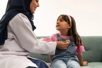 بورتريه مقرب لطبيبة عربية خليجية سعودية محجبة ترتدي المعطف الطبي الابيض و عباءة زرقاء تقوم بسماع نبضات قلب صغيرة ,طفلة عربية خليجية سعودية ترتدي الزي الكاجوال جالسة مطمئنة على اريكة العيادة , خلفية بيضاء