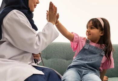 The profession of medicine is the noblest profession. An Arab Gulf Saudi female doctor wearing a hijab, an abaya, and a white medical coat is shaking hands with a little girl high-fiving. The little girl, an Arab Saudi Gulf child, is wearing casual attire and sitting next to the doctor. It is a close-up portrait with a white background.