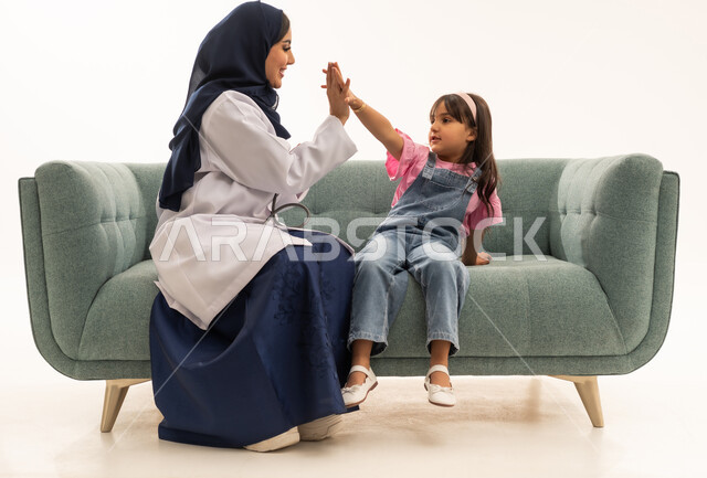 Commitment to work while maintaining fun and pleasant moments between doctors and patients, a full-body portrait of a veiled Saudi Gulf Arab female doctor wearing a white medical coat sitting on a sofa shaking hands with a little girl, a Saudi Gulf Arab girl wearing casual attire playing with the doctor shaking hands after work hours, white background.