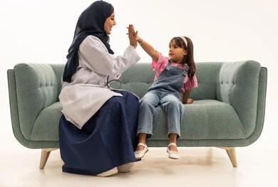 Commitment to work while maintaining fun and pleasant moments between doctors and patients, a full-body portrait of a veiled Saudi Gulf Arab female doctor wearing a white medical coat sitting on a sofa shaking hands with a little girl, a Saudi Gulf Arab girl wearing casual attire playing with the doctor shaking hands after work hours, white background.