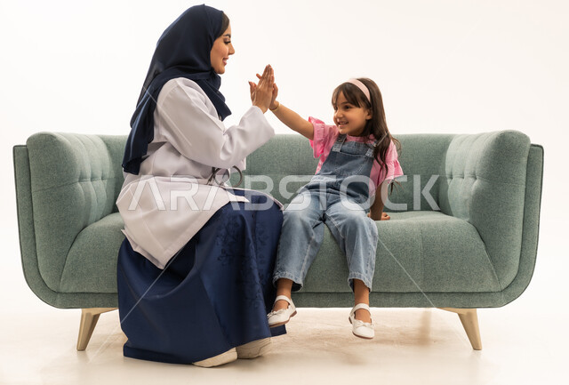 علاقة المحبة و المودة بين الاطباء و المرضى ,طبيبة عربية خليجية سعودية محجبة ترتدي عباءة زرقاء و المعطف الطبي الابيض تجلس على الاريكة بجانب الطفلة ,طفلة عربية خليجية سعودية ترتدي الزي الكاجوال تصافح الطبيبة بالطفين بمرح و سعادة , بورتريه خلفية بيضاء , الجسم بالطول الكامل