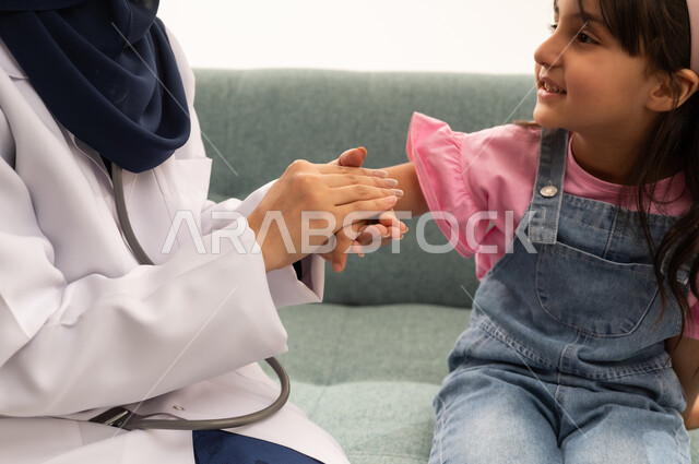 بورتريه مقرب ملئ بايماءات السعادة و السرور , طفلة عربية خليجية سعودية ترتدي الزي الكاجوال تنظر مبتسمة الى الطبيبة , طبيبة عربية خليجية سعودية محجبة تمسك يد الطفلة الصغيرة ,خلفية بيضاء