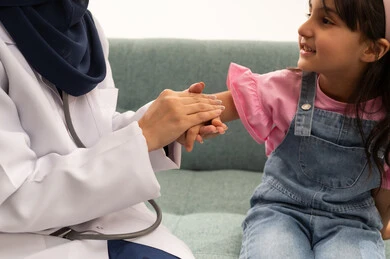 بورتريه مقرب ملئ بايماءات السعادة و السرور , طفلة عربية خليجية سعودية ترتدي الزي الكاجوال تنظر مبتسمة الى الطبيبة , طبيبة عربية خليجية سعودية محجبة تمسك يد الطفلة الصغيرة ,خلفية بيضاء