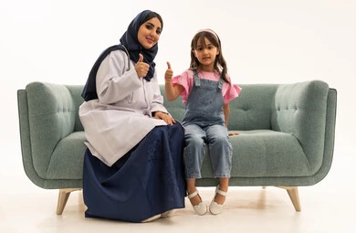 رفع اصبع الابهام للاعلى اشارة على جودة الخدمات المقدمة في هيئة الهلال الاحمر السعودي , طبيبة عربية خليجية سعودية محجبة ترتدي عباءة زرقاء تجلس على اريكة رمادية اللون , طفلة عربية خليجية سعودية ترتدي الزي الكاجوال تجلس بجانب الطبيبة المميزة ,ايماءات السعادة و السرور , بورتريه خلفية بيضاء ,الجسم بالطول الكامل