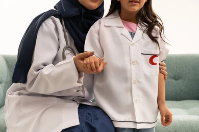 A Saudi Arabian Gulf Arab female doctor wearing a white medical coat and a hijab is holding the hand of a small girl. The girl, also a Saudi Arabian Gulf Arab, is dressed in a little doctor's outfit and is standing next to the doctor. This is a close-up portrait with a white background.