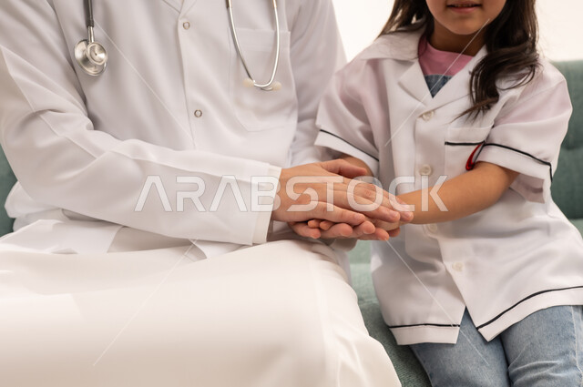 The relationship of love and affection between the doctors of the Saudi Red Crescent Authority and the patients, a Gulf Arab Saudi girl wearing a little doctor's outfit holding her doctor's hand safely, an Arab Saudi Gulf doctor wearing a white medical coat gently patting the small hands, close-up portrait, white background.