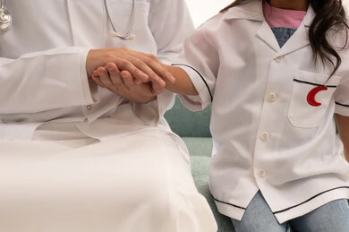 طفلة عربية خليجية سعودية ترتدي زي الطبيب الصغير  تمسك يد الطبيب , بورتريه مقرب لطبيب عربي خليجي سعودي يرتدي المعطفف الابيض الطبي يمسك اليد الصغيرة للاطمئنان على سلامتها , خلفية بيضاء