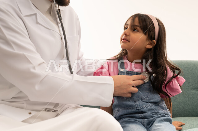 A Saudi Arabian Gulf Arab doctor wearing a white medical coat listens to the heartbeat of a little girl. The little girl, a Saudi Arabian Gulf Arab, is wearing casual attire and looking at the doctor during the medical examination. Close-up portrait, white background.