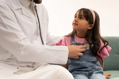 طبيب عربي خليجي سعودي يرتدي المعطف الطبي الابيض يستمع الى ضربات قلب الطفلة الصغيرة ,طفلة عربية خليجية سعودية ترتدي الزي الكاجوال تنظر الى الطبيب اثناء عملية الفحص الطبي , بورتريه مقرب ,خلفية بيضاء