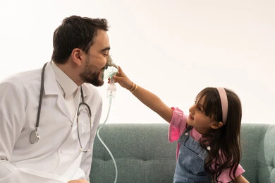 طفلة عربية خليجية سعودية صغيرة تداعب الطبيب بوضع قناع التنفس على الانف , طبيب عربي خليجي سعودي يرتدي المعطف الطبي الابيض يلعب مع الطفلة بعد انتهاء الفحص و الاطمئنان على الصحة , بورتريه مقرب خلفية بيضاء