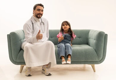 A small Arab Gulf Saudi girl wearing casual attire is sitting on a gray sofa next to a doctor. The Arab Gulf Saudi doctor is wearing a white medical coat and is raising his thumb as a sign of quality and mastery of work, in a full-body portrait with a white background.