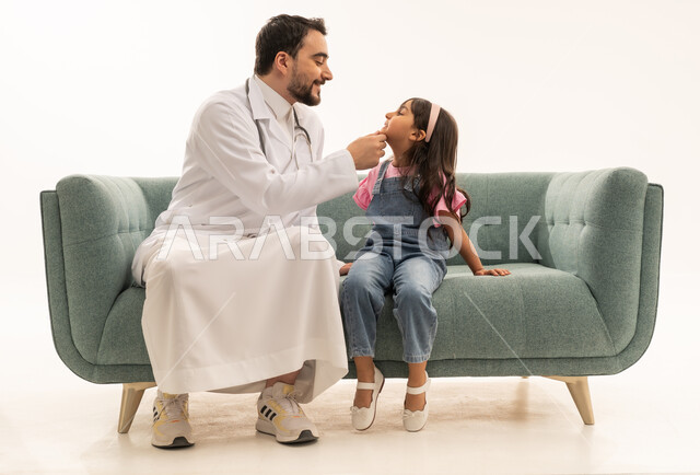 الفحص الدوري اللازم لحماية الاسنان ,طبيب عربي سعودي خليجي يرتدي المعطف الطبي الابيض اثناء فحص اللثة و الفم , طفلة عربية خليجية سعودية ترتدي الزي الاجوال تجلس على اريكة المستوصف ,بورتريه الجسم بالطول الكامل, خلفية بيضاء