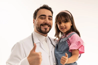 Looking at the camera with a thumbs up is a sign of quality and successful task completion. A close-up portrait of a Saudi Gulf Arab girl wearing casual attire, raising her thumb with gestures of happiness. An Arab Gulf Saudi doctor wearing a white medical coat is holding the little girl, with a white background.