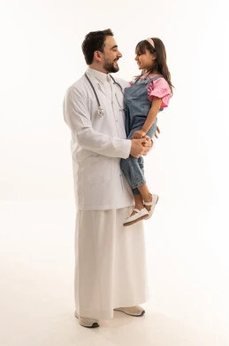 طبيب عربي خليجي سعودي يرتدي المعطف الطبي الابيض يحمل طفلة صغيرة وينظر اليها بابتسامة, طفلة عربية خليجية سعودية ترتدي الزي الكاجوال تنطر الى الطبيب بابتسامة أمان , بورتريه خلفية بيضاء ,الجسم بالطول الكامل
