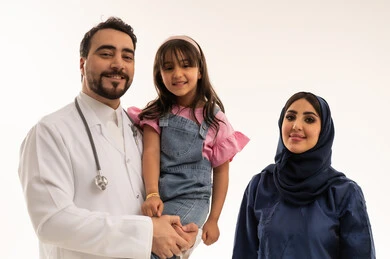 بورتريه مقرب لطبيب عربي خليجي سعودي يرتدي المعطف الابيض الطبي يحمل طفلة صغيرة عربية خليجية سعودية ترتدي الزي الكاجوال في سعادة ,امرأة عربية خليجية سعودية محجبة ترتدي عباءة زرقاء تقف بجانب الطبيب ,بورتريه مقرب ,خلفية بيضاء