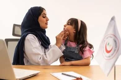 بورتريه مقرب لطبيبة  أسنان عربية خليجية سعودية محجبة ترتدي معطف طبي أبيض تمسك بفم الطلفة اثناء الكشف عن حالة الأسنان , طفلة علابية خليجية سعودية ترتدي الزي الكاجوال تجلس أمام الطبيبة بأمان و عدم خوف , خلفية بيضاء