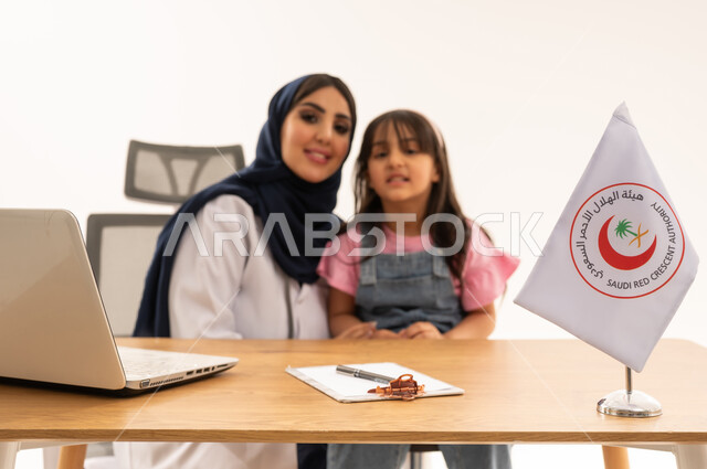 نظرة أمامية للكاميرا ,طبيبة عربية خليجية سعودية محجبة ترتدي المعطف الطبي الابيض تجلس بسعادة بعد الانتهاء من الفحص الطبي ,طفلة عربية سعودية خليجية صغيرة ترتدي الزي الكاجوال تجلس باطمئنان مع الطبيبة ,بورتريه مقرب , خلفية بيضاء