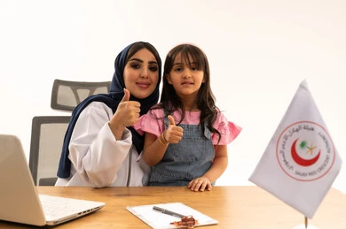 رفع اصبع الابهام اشارة على الجودة و السعادة بعد انتهاء الفحص الطبي,طبيبة عربية خليجية سعودية محجبة ترتدي المعطف الابيض تبتسم مع الطفلة الصغيرة ,بورتريه مقرب خلفية بيضاء