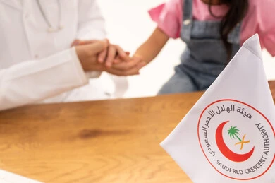 بورتريه مقرب , خلفية بيضاء ,طبيب عربي سعودي خليجي يرتدي المعطف الابيض يقوم باحتضان يد الفتاة الصغيرة بعد نهاية الفحص الطبي ,راية الهلال الاحمر السعودي رمز الصحة و الامان