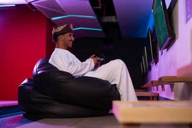 النظر بسعادة الي الشاشة المعلقة على الحائط , رجل عربي خليجي عماني يرتدي دشداشة بيضاء و عمامة يجلس على مقعد اسود اسفنجي , ايماءات السرور لقضاء الاوقات الممتعة