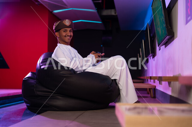 شاب عربي خليجي عماني يرتدي الدشداشة البيضاء و العمامة البنية يجلس على المقعد الاسود الاسفنجي , النظر للكاميرا بابتسامة , الامساك بجهاز التحكم عن بعد الخاص بالالعاب الالكترونية