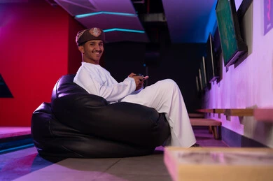 شاب عربي خليجي عماني يرتدي الدشداشة البيضاء و العمامة البنية يجلس على المقعد الاسود الاسفنجي , النظر للكاميرا بابتسامة , الامساك بجهاز التحكم عن بعد الخاص بالالعاب الالكترونية