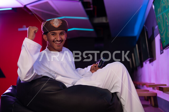 Raising a fist in the air is a sign of celebrating victory. An Arab Gulf Omani man wearing a brown turban and a white dishdasha looks at the camera with happiness and joy, sitting on a black sponge seat, with gestures of delight.