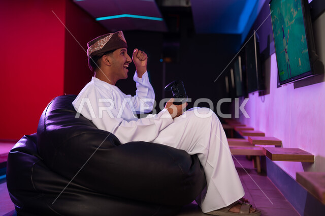 تعابير البهجة و الاحتفال بالفوز في اللعبة الالكترونية , رجل عربي خليجي عماني يرتدي عمامة بنية و دشداشة بيضاء يرفع قبضة اليد في الهواء علامة على تحقيق النصر , ايماءات الفرحة و السرور , النظر الي الشاشة المعلقة على الحائط