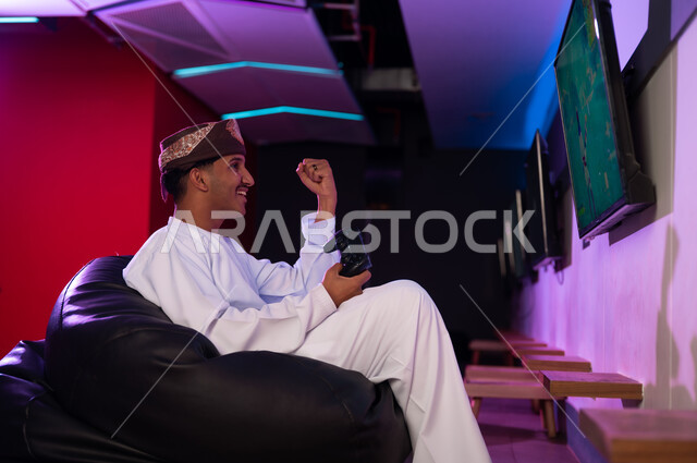 التلويح بقبضة اليد في الهواء علامة الاحتفال بالفوز , رجل عربي خليجي عماني يرتدي الدشداشة البيضاء و العمامة البنية يحمل جهاز التحكم عن بعد الخاص بالالعاب الالكترونية , ايماءات البهجة و السعادة