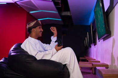 التلويح بقبضة اليد في الهواء علامة الاحتفال بالفوز , رجل عربي خليجي عماني يرتدي الدشداشة البيضاء و العمامة البنية يحمل جهاز التحكم عن بعد الخاص بالالعاب الالكترونية , ايماءات البهجة و السعادة