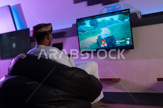 الجلوس على المقعد الاسود الاسفنجي المريح اثناء اللعب داخل مركز الالعاب الالكترونية , رجل عربي خليجي عماني يرتدي الدشداشة البيضاء و العمامة يحمل جهاز التحكم عن بعد , النظر الي الشاشة المعلقة على الحائط